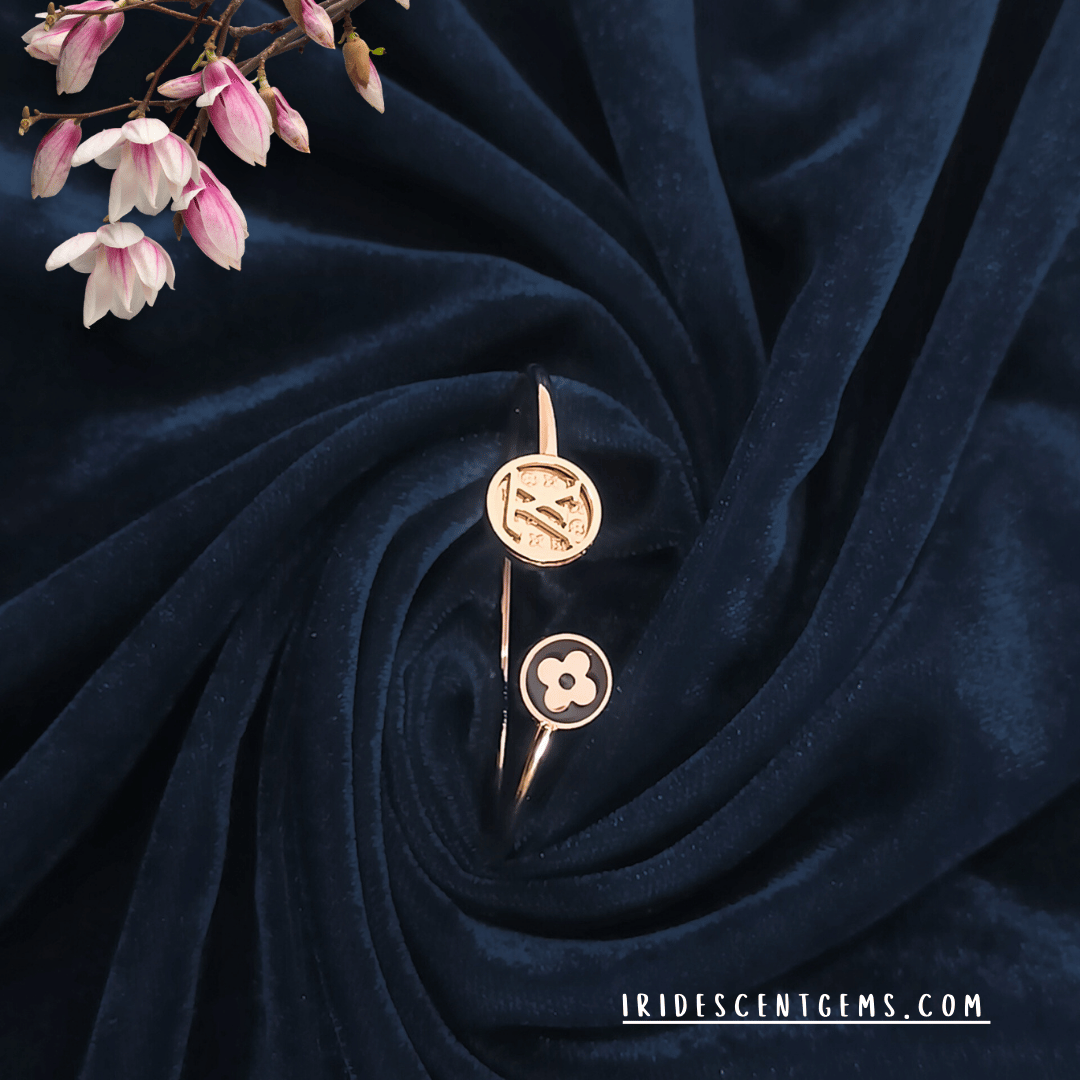 A stylish open cuff bracelet in gold with modern emblematic designs, elegantly displayed on a deep navy velvet background with soft pink floral accents.