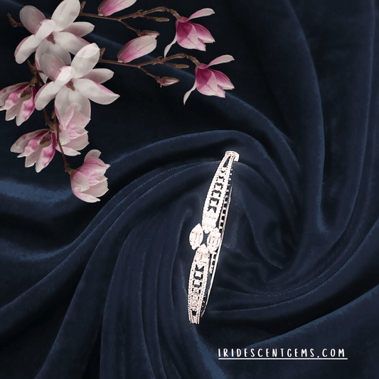 A dazzling diamond-studded bracelet with intricate detailing, elegantly placed on a deep navy velvet fabric, accompanied by delicate pink and white blossoms. A statement of luxury and grace by Iridescent Gems.
