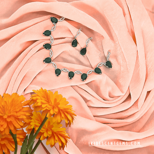 A luxurious silver necklace set adorned with teardrop-shaped emerald-green gemstones, paired with matching earrings, elegantly displayed on soft peach fabric with vibrant marigold flowers.
