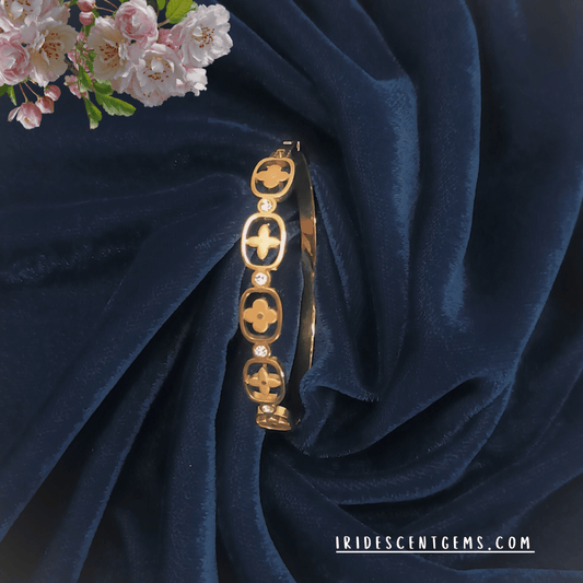 A sophisticated gold-plated bracelet featuring intricate floral cutout patterns and sparkling stones, elegantly displayed on a deep navy velvet fabric with soft pink blossoms. A statement piece from Iridescent Gems.