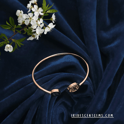 A sleek and contemporary gold bracelet featuring intricate motifs, resting gracefully on plush dark blue fabric, accompanied by delicate blossoms.