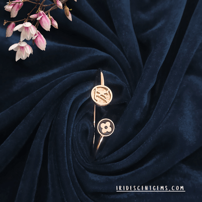 A stylish open cuff bracelet in gold with modern emblematic designs, elegantly displayed on a deep navy velvet background with soft pink floral accents.