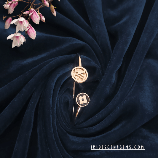 A stylish open cuff bracelet in gold with modern emblematic designs, elegantly displayed on a deep navy velvet background with soft pink floral accents.