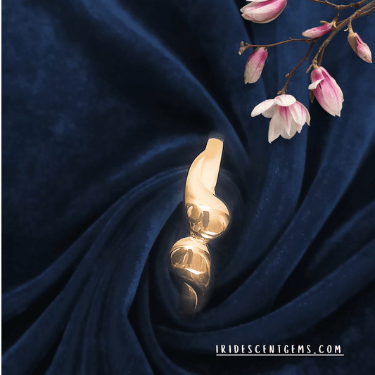 A modern gold-plated bracelet with a sleek, twisted design, elegantly displayed on a deep navy velvet fabric with delicate pink blossoms. A perfect blend of sophistication and minimalism from Iridescent Gems.