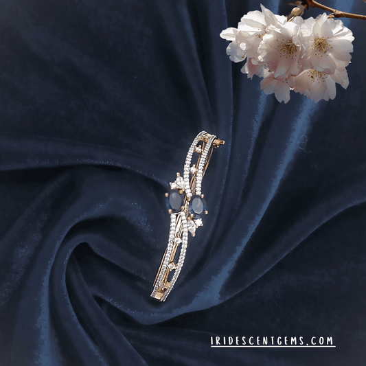 A stunning gold-toned bracelet adorned with deep blue sapphire-like gemstones and delicate crystal accents, set against a rich navy velvet background with white blossoms.