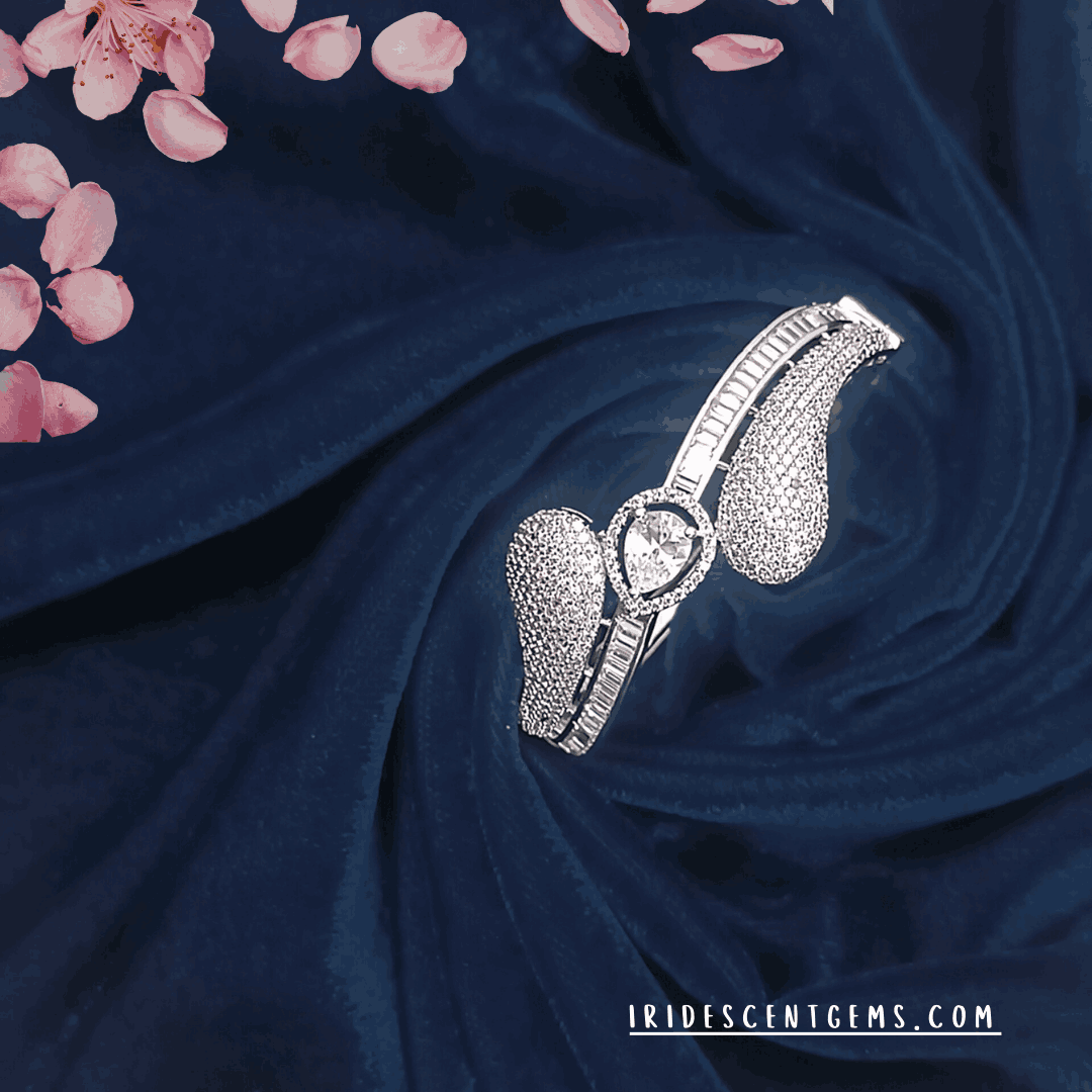 An exquisite silver bracelet featuring a teardrop-shaped diamond at the center, surrounded by intricate stone detailing, elegantly displayed on deep navy velvet with soft pink floral petals.